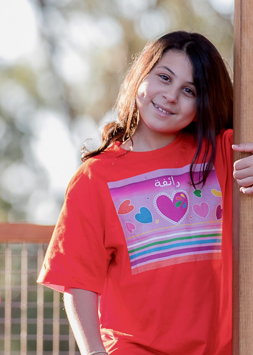 Our stunning model 'Raifa' is wearing the Jewels in My Heart Haluna Happy Names T-shirt in Orange with an Arabic personalisation. She is looking straight at the camera and smiling. She is at the park, with the evening sunlight falling on her dark hair and right cheek.