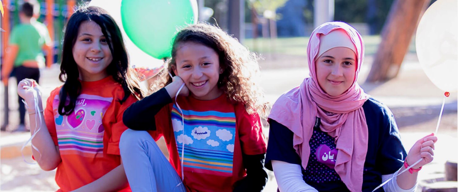T-shirts bearing Haluna Happy Names exclusive colourful personalised designs with hearts, rainbows and clouds with fun Arabic fonts are worn by three smiling girls with one in hijab in a park