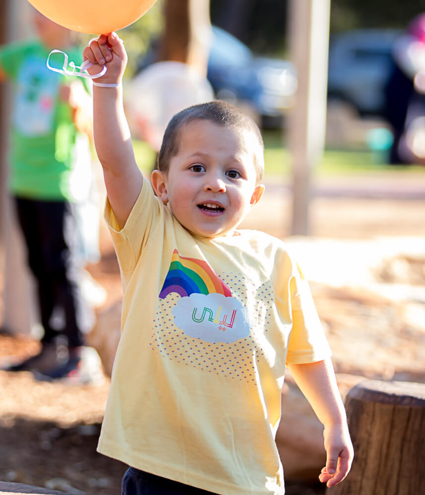 Sunlight falls on the side of the face of an adorable short-haired brown-eyed boy. He raises his arm holding a balloon, wearing a Haluna Happy Names 'End of the Rainbow' personalised yellow toddler t-shirt bearing the name 'Layla' in Arabic.