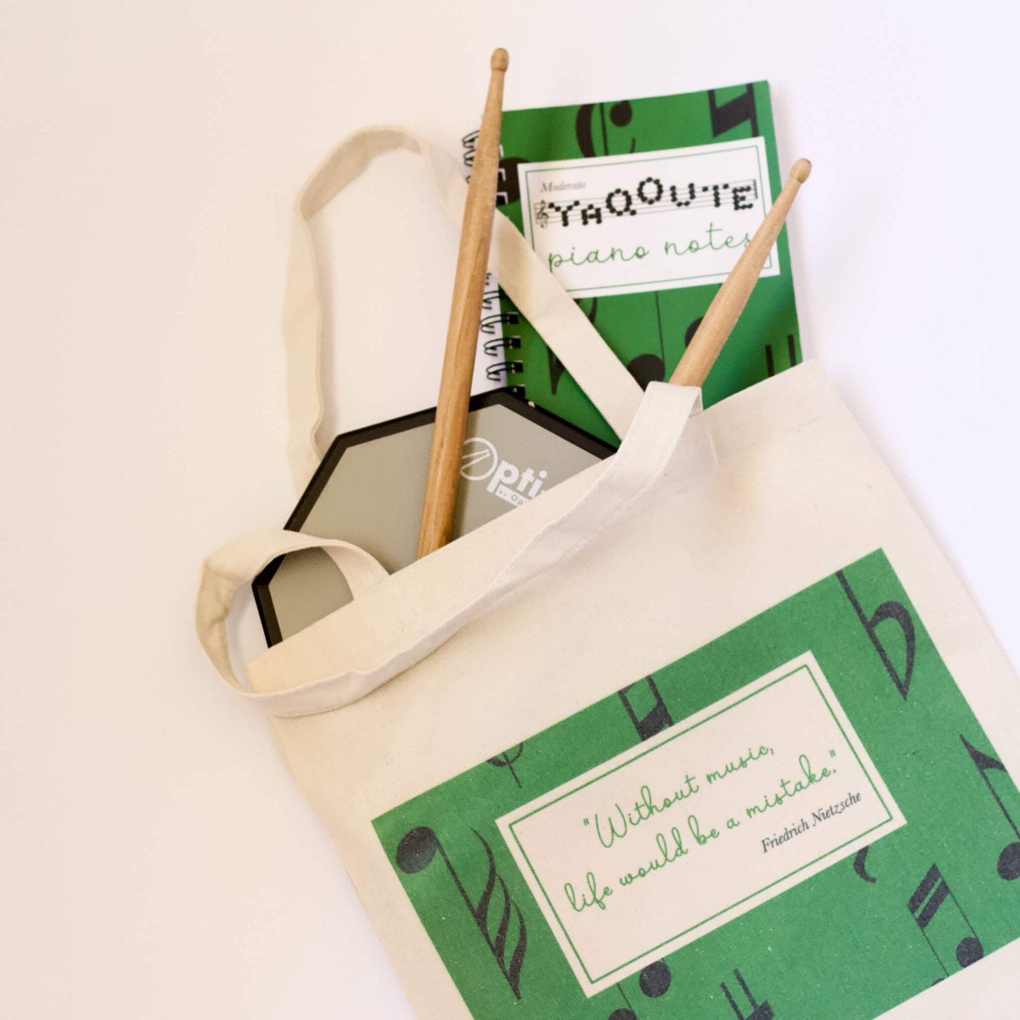The Haluna Happy Names Musically range of bags, notebooks, ID tags, mugs and more is perfect for budding and experienced musicians alike. Forest green bag with Nietzsche quote, matching notebook shown here in flatlay with drumsticks and drum practice pad.