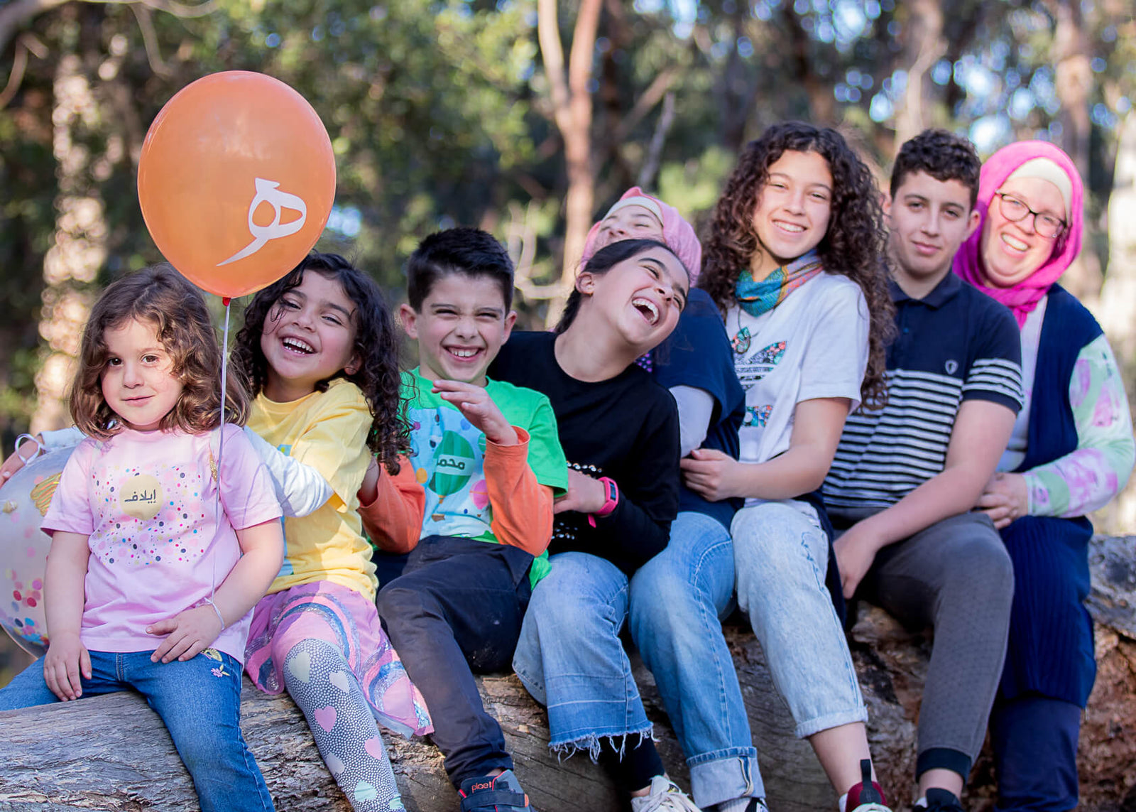 Helen the founder of Haluna Happy Names sits on a log behind her seven children lined from eldest to youngest. The youngest five are wearing colourful t-shirts personalised with their names in Arabic. They are all laughing and smiling in afternoon sun. 