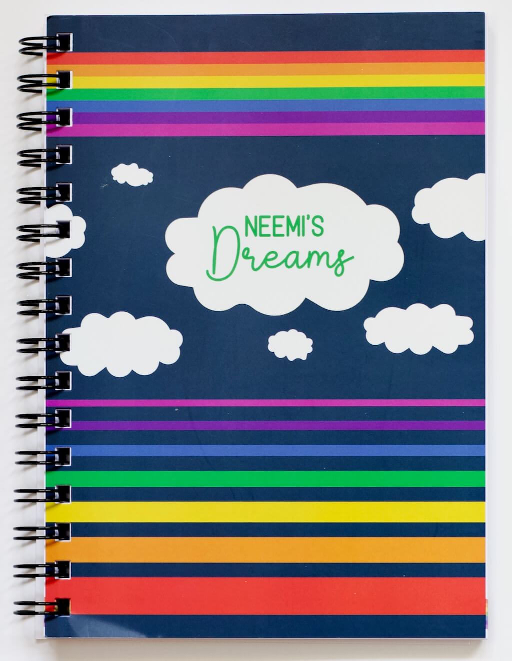 Cover of Haluna Happy Names &#39;In the Sky&#39; spiral-bound personalised A5 notebook in &#39;Night&#39;, showing from top to bottom a band of rainbow stripes, a flotilla of fluffy clouds, the biggest of which bears the notebook&#39;s title &quot;Neemi&#39;s Dreams&quot;, and then separated set of rainbow stripes in inverse order, gradually getting wider from violet to red. The rainbows and clouds are against a dark navy night sky background.