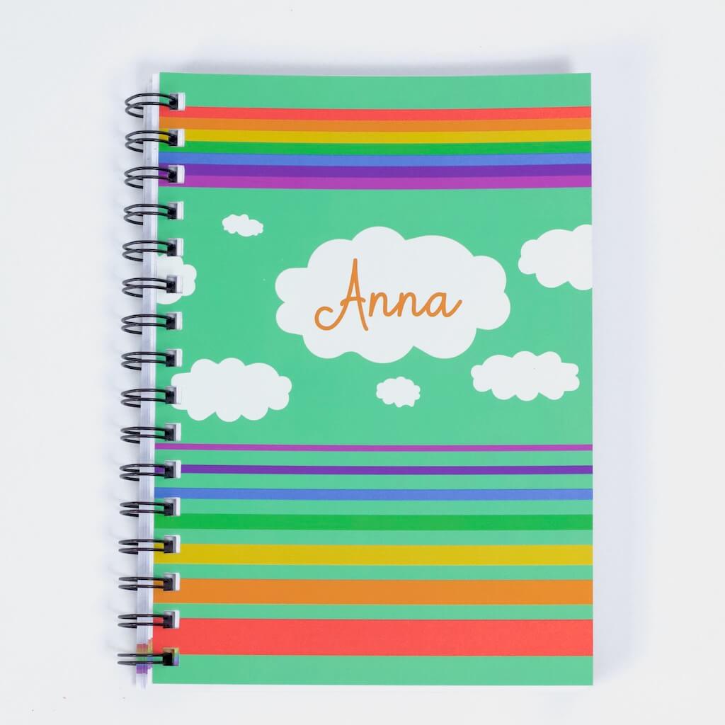Cover of Haluna Happy Names &#39;In the Sky&#39; spiral-bound personalised A5 notebook in &#39;Storm&#39;, showing from top to bottom a band of rainbow stripes, a flotilla of fluffy clouds, the biggest of which bears the notebook&#39;s owner&#39;s name, &quot;Anna&quot;, and then separated set of rainbow stripes in inverse order, gradually getting wider from violet to red. The rainbows and clouds are against an electric storm green background.