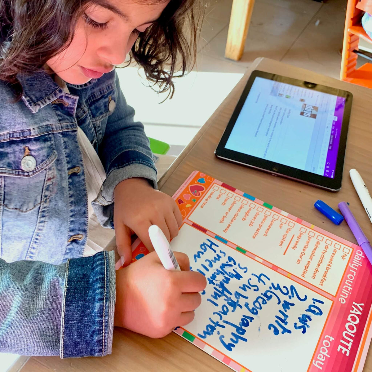 A young girl with dark hair writes a list of to-dos for today in blue on her Raspberry personalised &#39;Jewels In My Heart&#39; design Haluna Happy Names whiteboard with routine tracker panel.