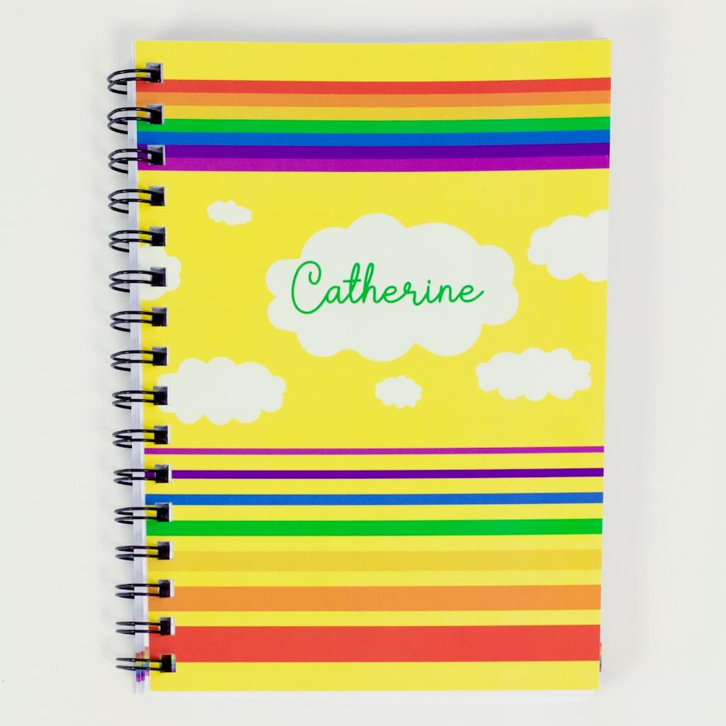 Cover of Haluna Happy Names &#39;In the Sky&#39; spiral-bound personalised A5 notebook in &#39;Sunshine&#39;, showing from top to bottom a band of rainbow stripes, a flotilla of fluffy clouds, the biggest of which bears the notebook&#39;s owner&#39;s name, &quot;Catherine&quot;, and then separated set of rainbow stripes in inverse order, gradually getting wider from violet to red. The rainbows and clouds are against a bright sunshine yellow background.