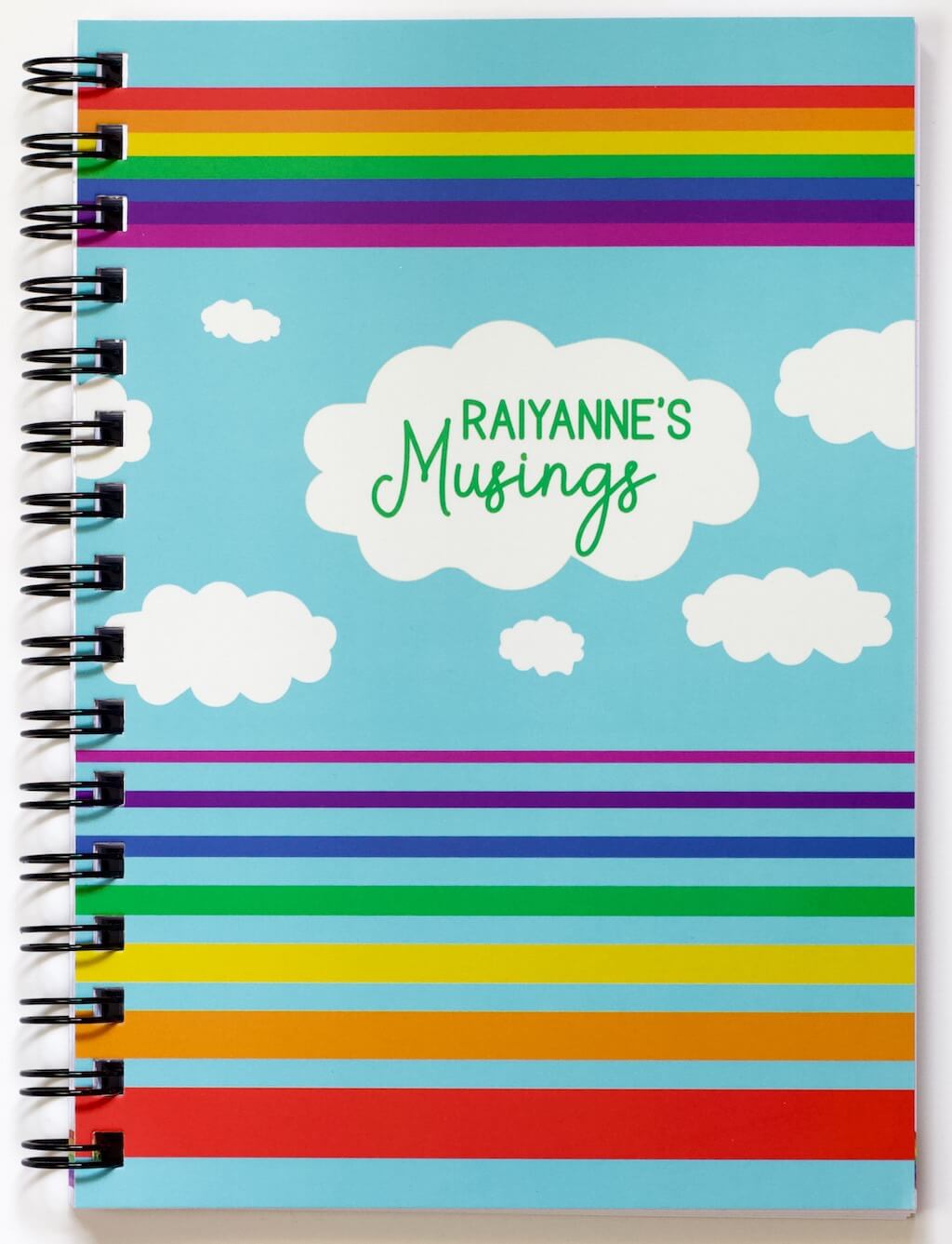 Cover of Haluna Happy Names &#39;In the Sky&#39; spiral-bound personalised A5 notebook in &#39;Day&#39;, showing from top to bottom a band of rainbow stripes, a flotilla of fluffy clouds, the biggest of which bears the notebook&#39;s title &quot;Raiyanne&#39;s Musings&quot;, and then separated set of rainbow stripes in inverse order, gradually getting wider from violet to red. The rainbows and clouds are against a daytime blue sky background.