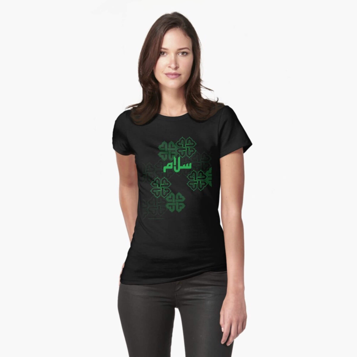 A white female model with hazel eyes and mid-brown shoulder length hair wears the Haluna Happy Names exclusive &#39;Salaam—Arabic Key Words&#39; design black fitted t-shirt and a pair of tight black jeans. The word &#39;salaam&#39; is written in green in Arabic kufic-style font across the chest of the model against a complementary green patterned background print. The image cuts off mid-thigh. 