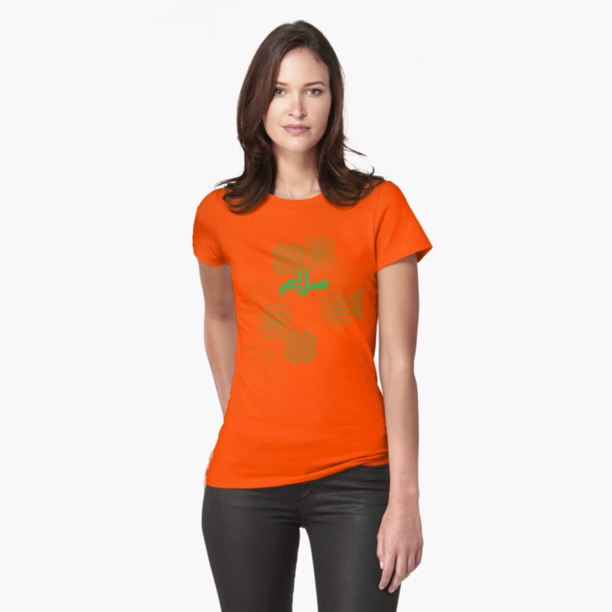 A white female model with hazel eyes and mid-brown shoulder length hair wears the Haluna Happy Names exclusive &#39;Salaam—Arabic Key Words&#39; design bright orange loosely fitted t-shirt and a pair of tight black jeans. The word &#39;salaam&#39; is written on the tshirt in green in Arabic using a kufic-style font, across the chest of the model against a complementary green patterned background print. The tshirt ends at the woman&#39;s hips and the image cuts off mid-thigh.