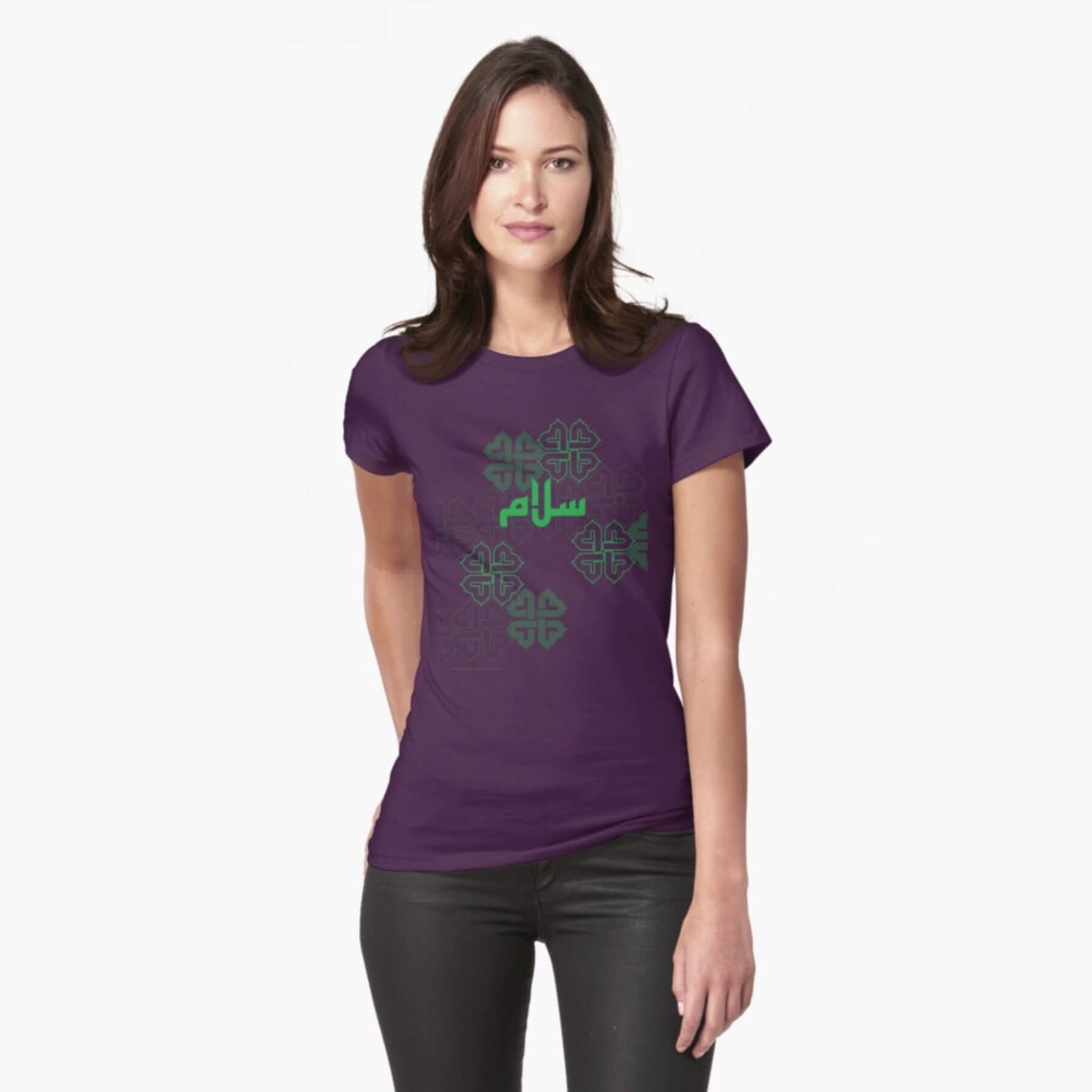 A white female model with hazel eyes and mid-brown shoulder length hair wears the Haluna Happy Names exclusive 'Salaam—Arabic Key Words' design purple fitted t-shirt and a pair of tight black jeans. The word 'salaam' is written in green in Arabic kufic-style font against a complementary green patterned background. The image cuts off mid-thigh. 