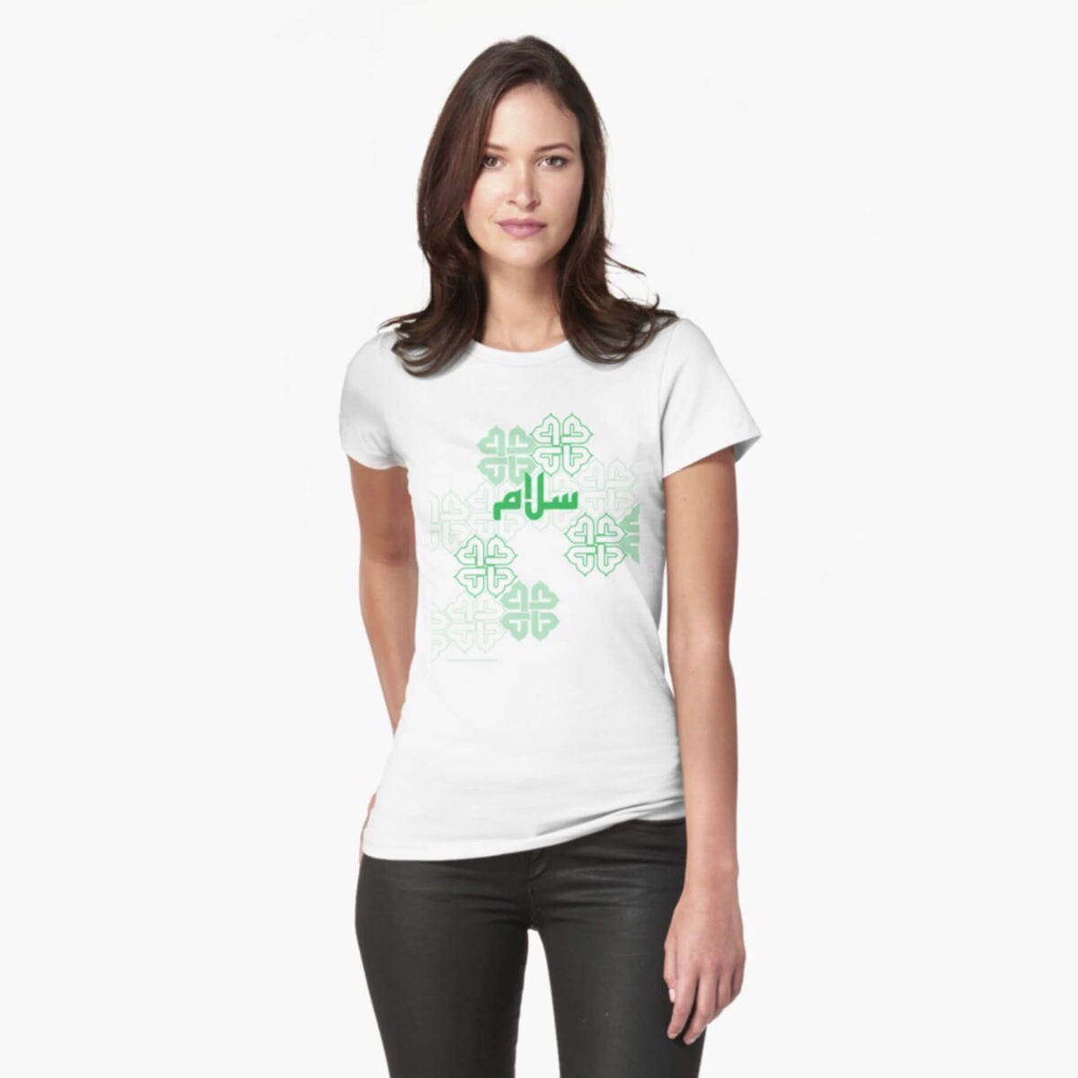 A white female model with hazel eyes and mid-brown shoulder length hair wears the Haluna Happy Names exclusive &#39;Salaam—Arabic Key Words&#39; design white fitted t-shirt and a pair of tight black jeans. The word &#39;salaam&#39; is written on the tshirt in green in Arabic using a kufic-style font, across the chest of the model against a complementary green patterned background print. The tshirt ends at the woman&#39;s hips and the image cuts off mid-thigh. 