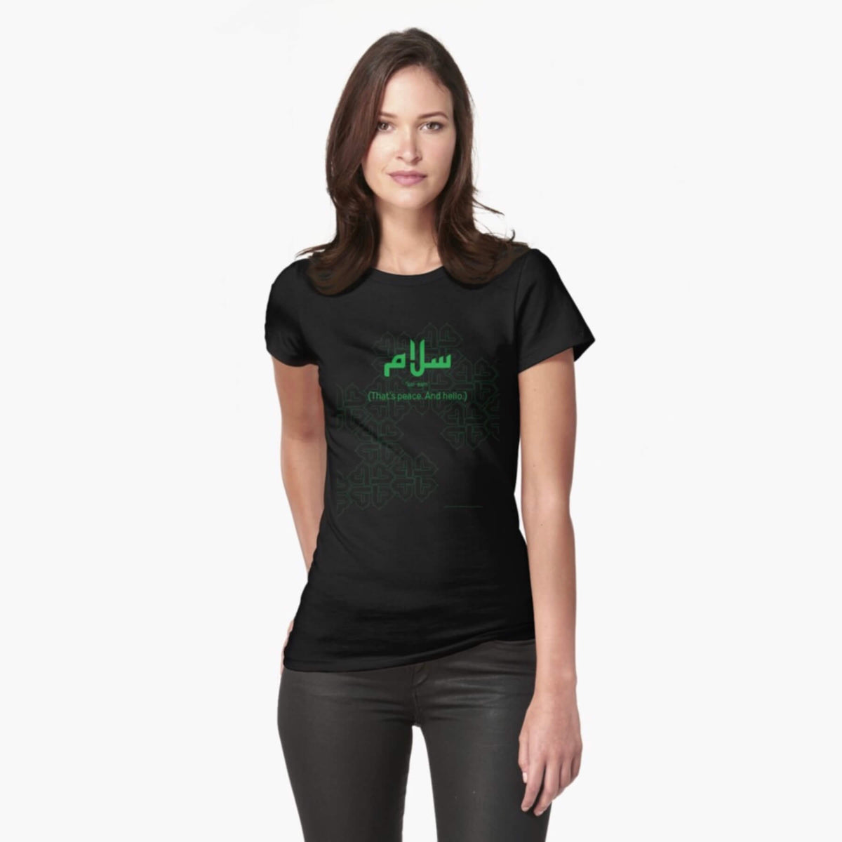 A white female model with hazel eyes and brown shoulder-length hair wears the Haluna Happy Names exclusive &#39;Salaam—Arabic Key Words&#39; design black fitted t-shirt and a pair of tight black jeans. The word &#39;salaam&#39; is written in green in Arabic with a kufic-style font, and beneath it in English the words &quot;Sal-aam&quot; and beneath that the words &#39;(That means peace. And hello)&#39;. The text is positioned on the tshirt so it falls across the chest of the model against a complementary green patterned background print. 