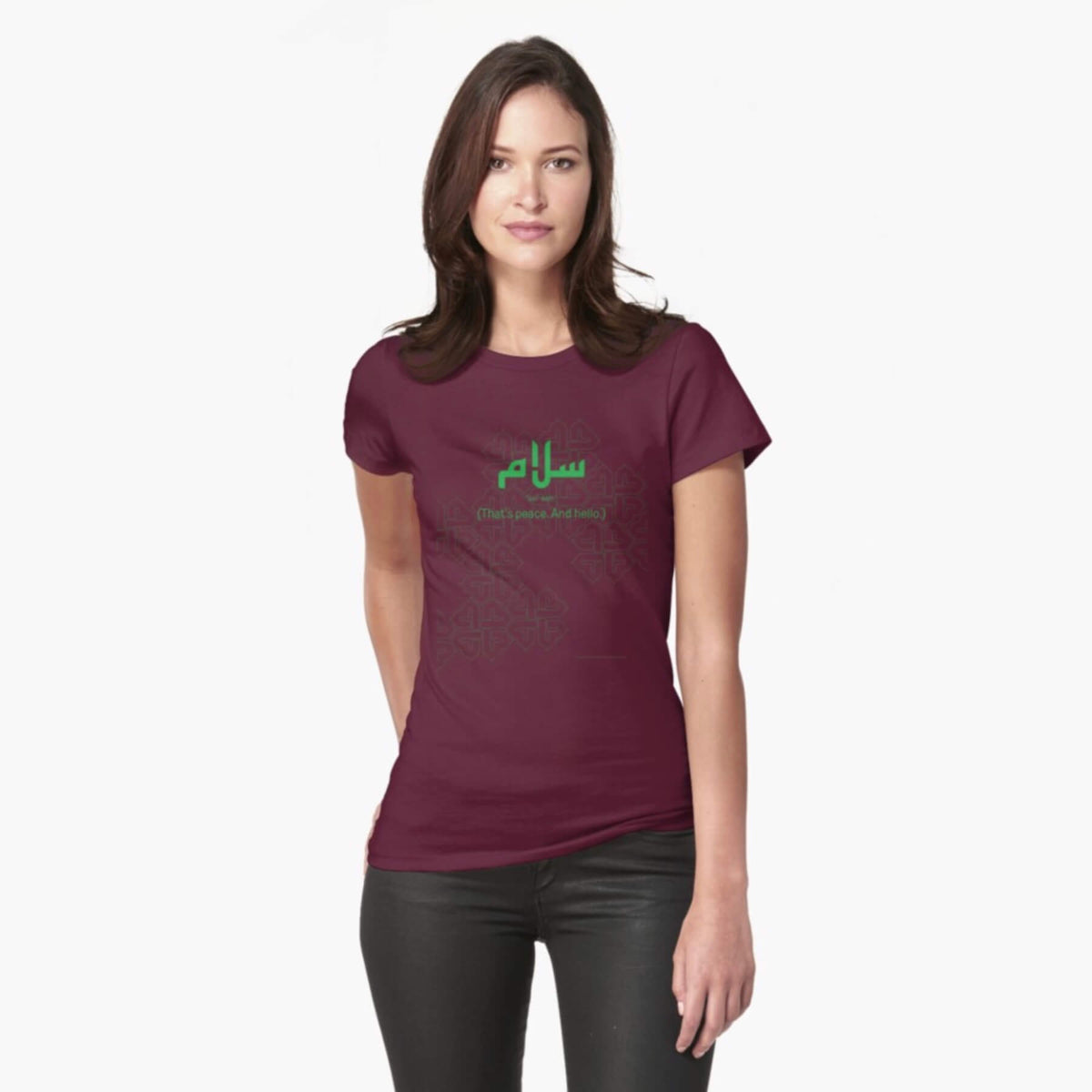 A white female model with hazel eyes and brown shoulder-length hair wears the Haluna Happy Names exclusive &#39;Salaam—Arabic Key Words&#39; design burgundy fitted t-shirt and a pair of tight black jeans. The word &#39;salaam&#39; is written in green in Arabic with a kufic-style font, and beneath it in English the words &quot;Sal-aam&quot; and beneath that the words &#39;(That means peace. And hello)&#39;. The text is positioned on the tshirt so it falls across the chest of the model against a complementary green patterned background print.