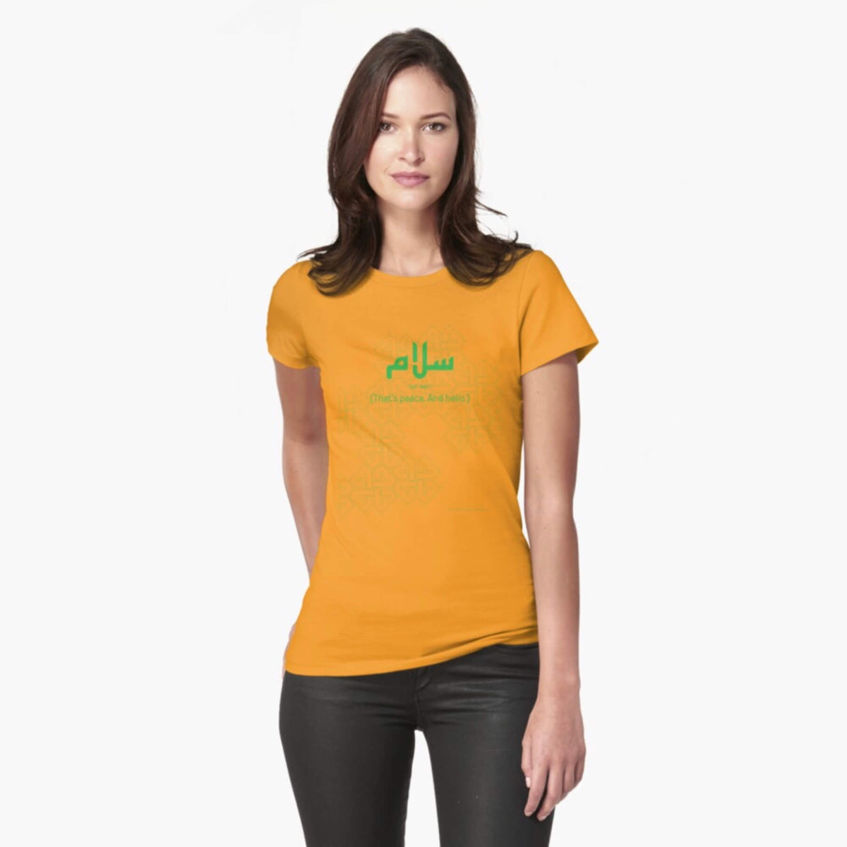 A white female model with hazel eyes and brown shoulder-length hair wears the Haluna Happy Names exclusive &#39;Salaam—Arabic Key Words&#39; design gold fitted t-shirt and a pair of tight black jeans. The word &#39;salaam&#39; is written in green in Arabic with a kufic-style font, and beneath it in English the words &quot;Sal-aam&quot; and beneath that the words &#39;(That means peace. And hello)&#39;. The text is positioned on the tshirt so it falls across the chest of the model against a complementary green patterned background print.