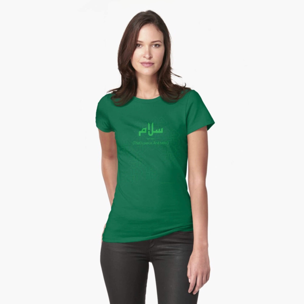 A white female model with hazel eyes and brown shoulder-length hair wears the Haluna Happy Names exclusive &#39;Salaam—Arabic Key Words&#39; design emerald-green fitted t-shirt and tight black jeans. The word &#39;salaam&#39; is written in green in Arabic with a kufic-style font, and beneath it in English the words &quot;Sal-aam&quot; and beneath that the words &#39;(That means peace. And hello)&#39;. The text is positioned on the tshirt so it falls across the chest of the model against a complementary green patterned background print.