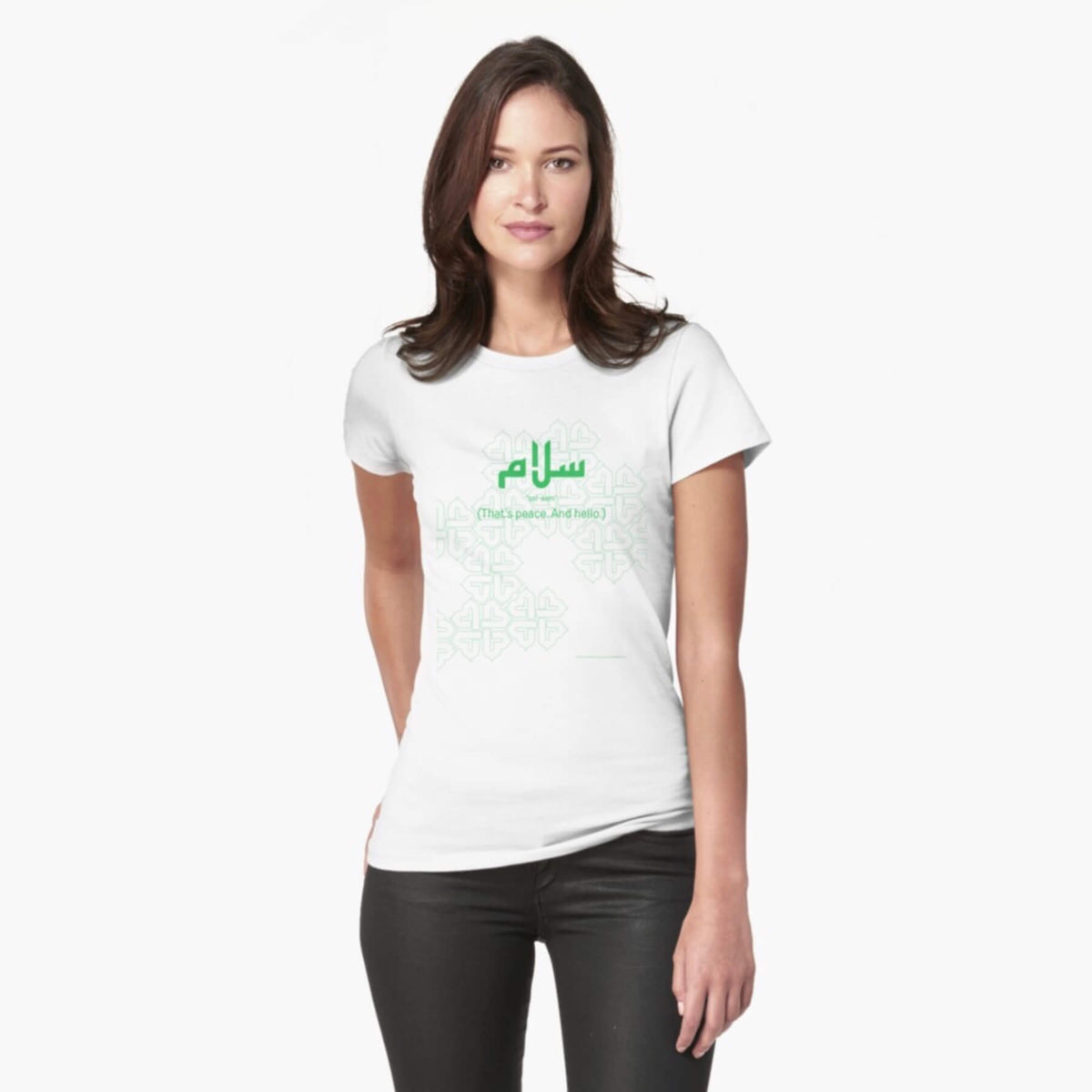 A white female model with hazel eyes and brown shoulder-length hair wears the Haluna Happy Names exclusive &#39;Salaam—Arabic Key Words&#39; design fitted t-shirt in white, and tight black jeans. The word &#39;salaam&#39; is written in green in Arabic with a kufic-style font, and beneath it in English the words &quot;Sal-aam&quot; and beneath that the words &#39;(That means peace. And hello)&#39;. The text is positioned on the tshirt so it falls across the chest of the model against a complementary green background pattern.