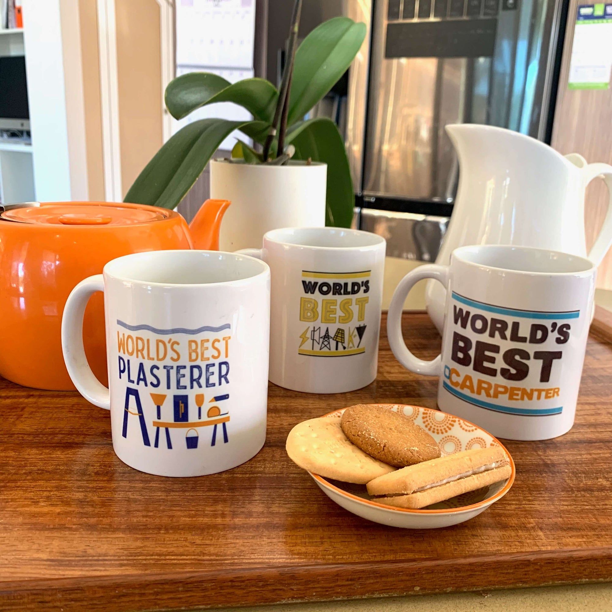 Exclusive Haluna Happy Names mug designs for the world's best plasterer, carpenter and sparky on a tray with orange teapot, white milk jug and of course a few biccies. Pot plant behind. Ready for a cuppa? These guys deserve one.