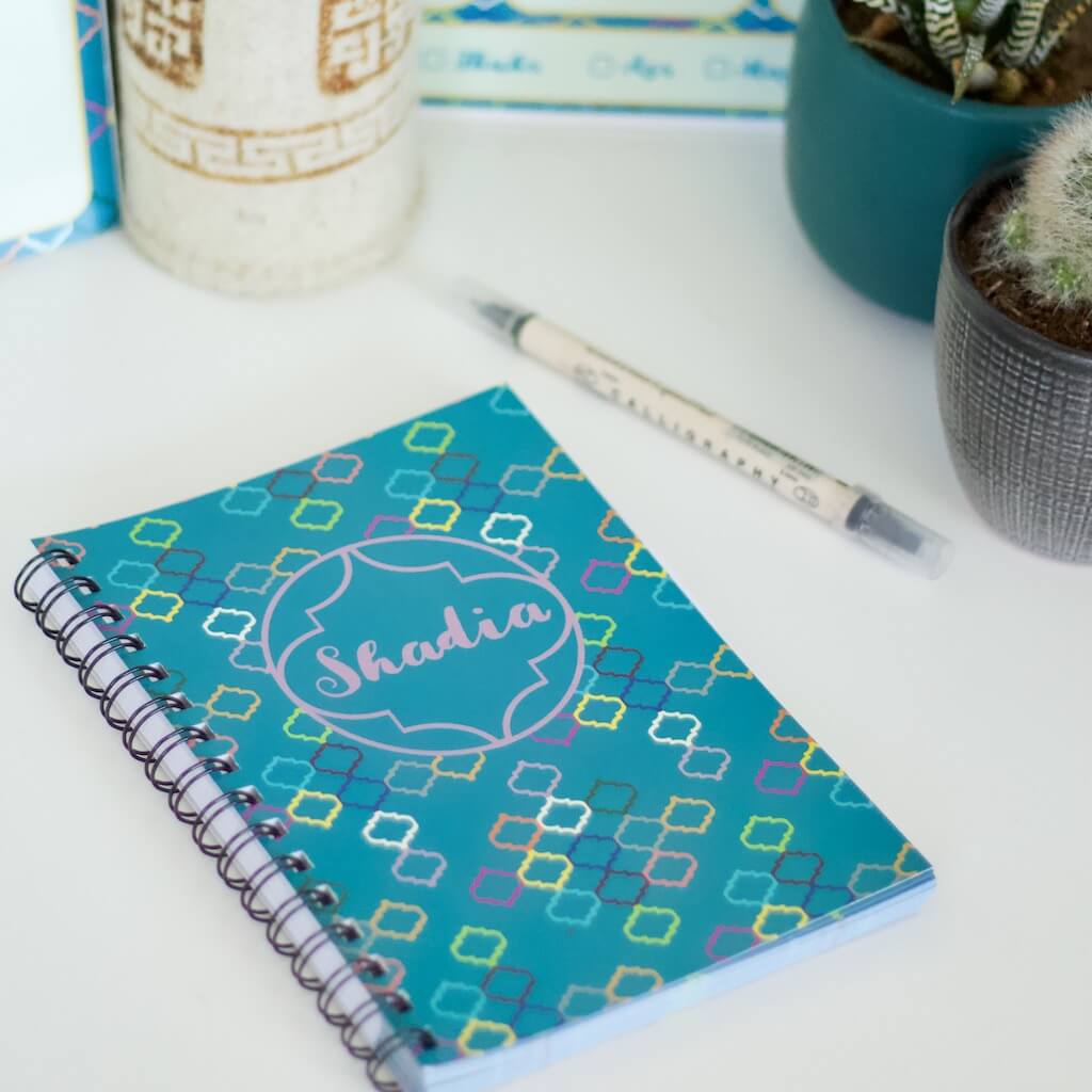 Gorgeous teal personalised &#39;Maroc Magic&#39; design notebook. Cover bears name in centre of Moroccan line motif against a background of repeated motifs in many colours. Lies on desk with matching Maroc Magic whiteboard and routine tracker in background with Mongolian felt pen pot, two cacti and a black pen